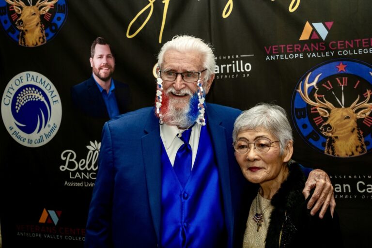 Veteran with a red, white and blue beard and his guest in front of the sponsor wall at the 2025 Military Ball